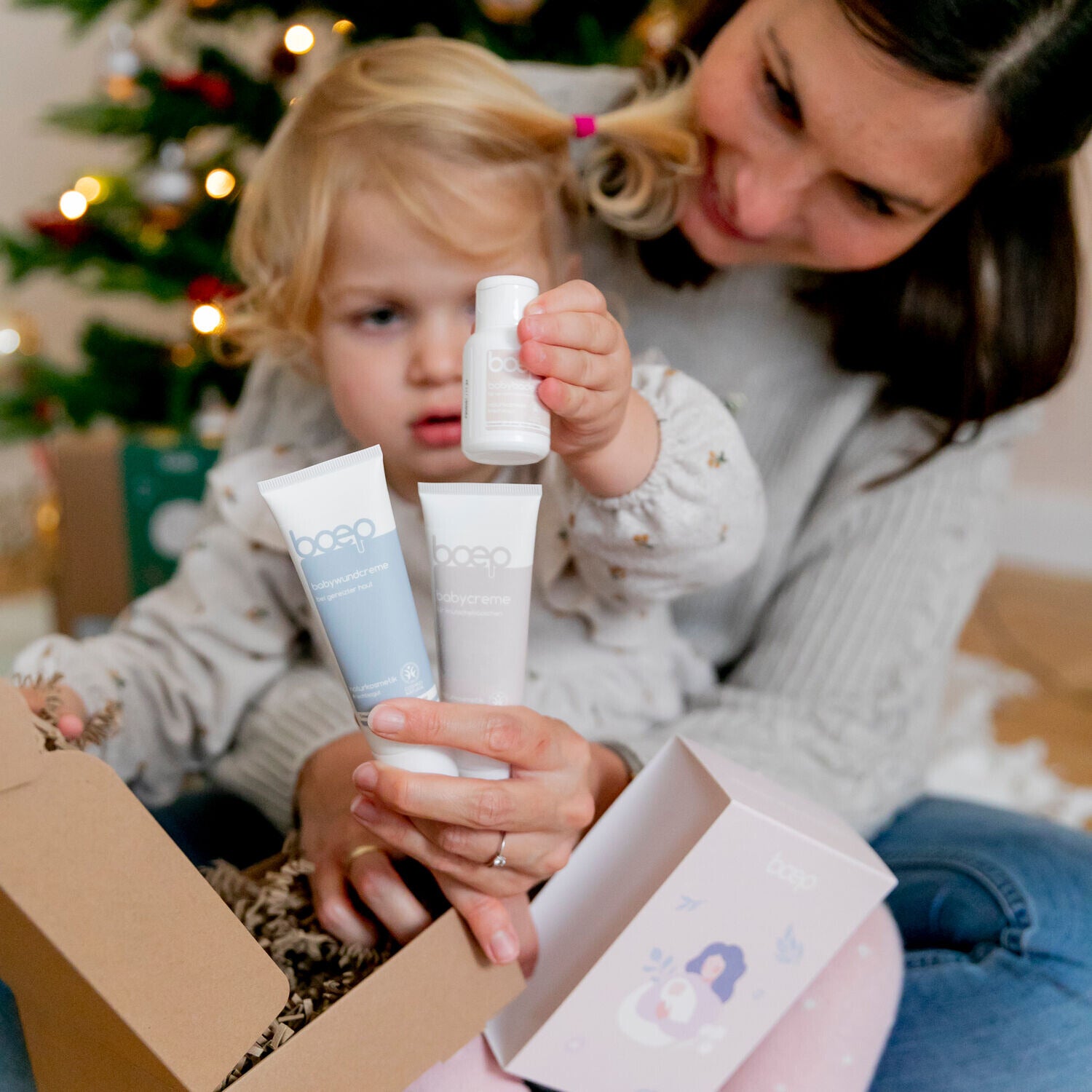 Mama und Kind packen das Geschenkset von das boep mit babywundcreme, babycreme und dem babymandelöl in Reisegröße vor dem Weihnachtsbaum aus