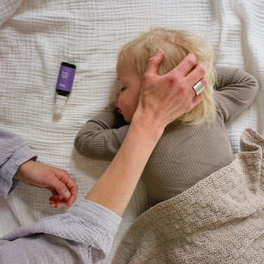 Das Gute Nacht Spray von das boep, das als Kissenspray oder als Raumspray angewendet werden kann, liegt auf einer Matratze neben einem schlafenden Kind und die Mutter hält die Hand an seinen Kopf.